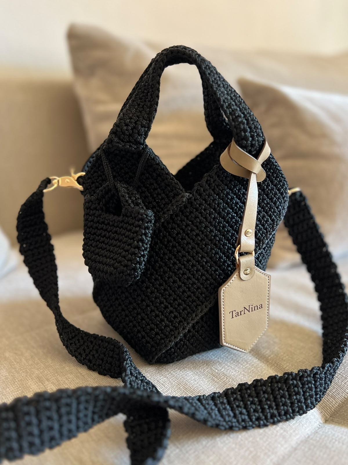 Black TarNina handcrafted crochet shoulder bag with beige leather tag, displayed on a neutral sofa