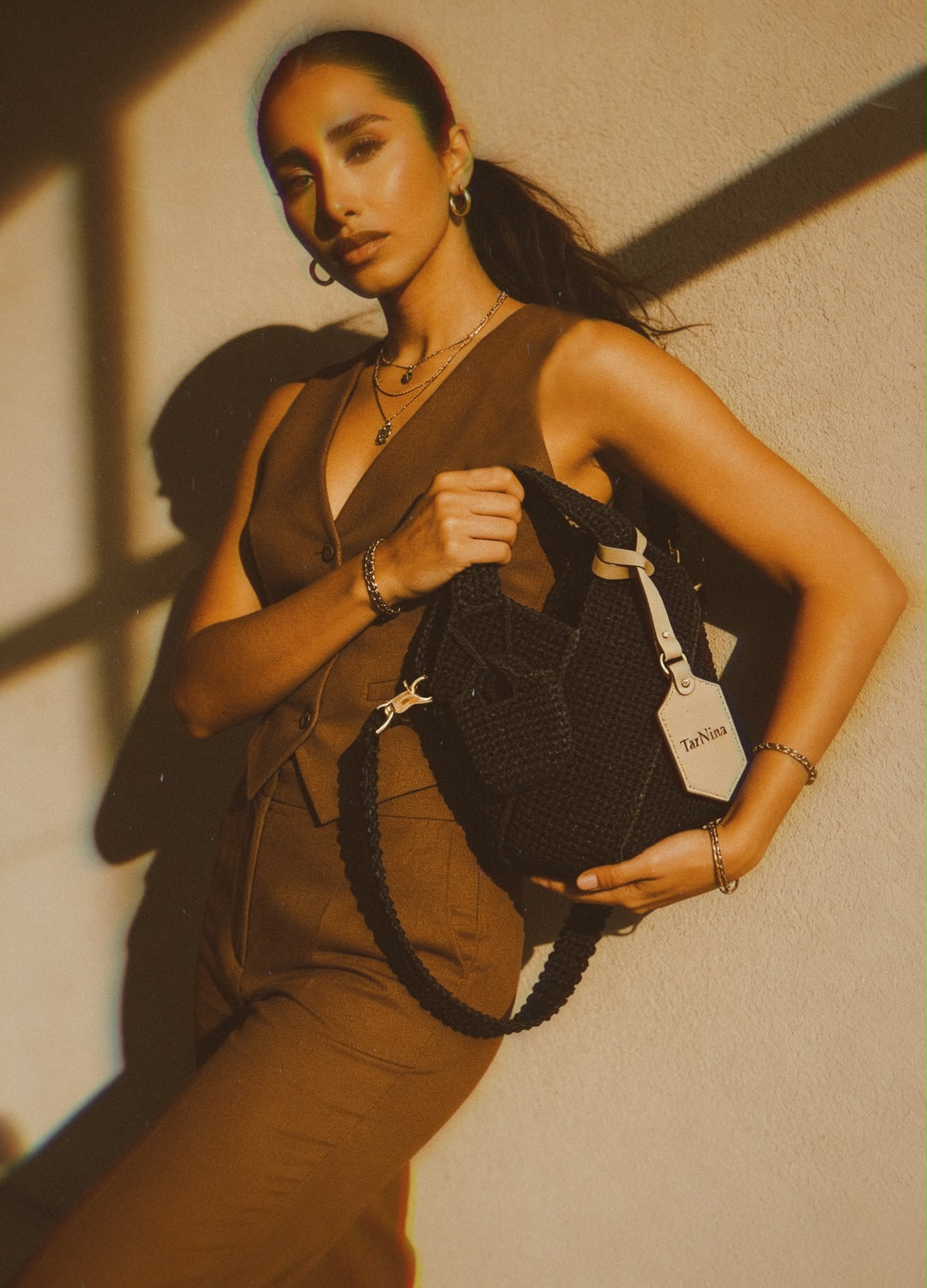 Nina holding a dark handcrafted TarNina crochet bag, standing against a warm textured wall with dramatic golden shadows — TarNina campaign